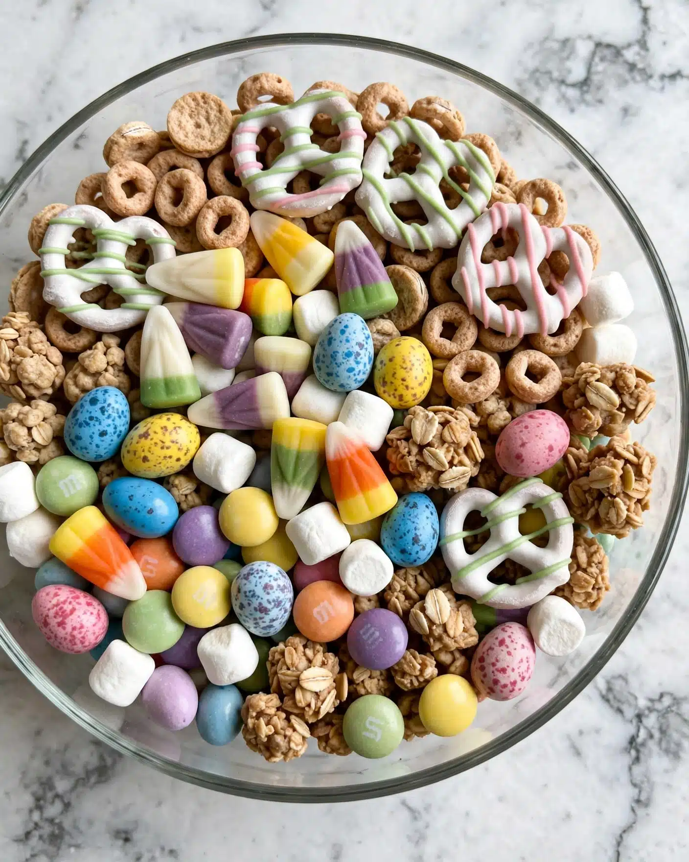 5 Minute Holiday Snack Mix Recipe 5 A clear glass bowl filled with layers of colorful candy and snacks sits on a white marbled surface. The top layer includes light brown circular cereal, white pretzels drizzled with green and pink stripes, pastel candy corns in white, yellow, green, and purple colors, and small white marshmallows. There are also speckled bright blue, yellow, and pink candy eggs, light brown clusters of cookies or cereal, and round candy-coated chocolates in pastel shades of yellow, green, purple, orange, and blue. All the items are arranged closely together, showing a mix of smooth, crunchy, and soft textures. photo taken with an iphone --ar 4:5 --v 7