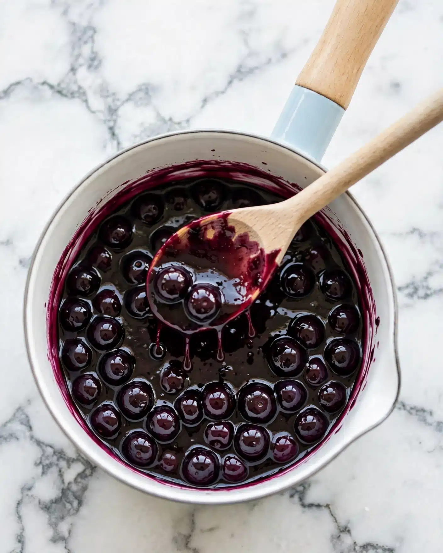 Vegan Blueberry Ghost Hand Pies Recipe 4 A white saucepan filled with a thick, dark purple sauce containing whole, shiny blueberries. A wooden spoon with light wood color is dipped into the sauce, resting inside the pan with some of the sauce dripping back into the pan. The saucepan has a wooden handle with a light blue section near the pan itself. The background surface is white with a marbled texture. Photo taken with an iphone --ar 4:5 --v 7